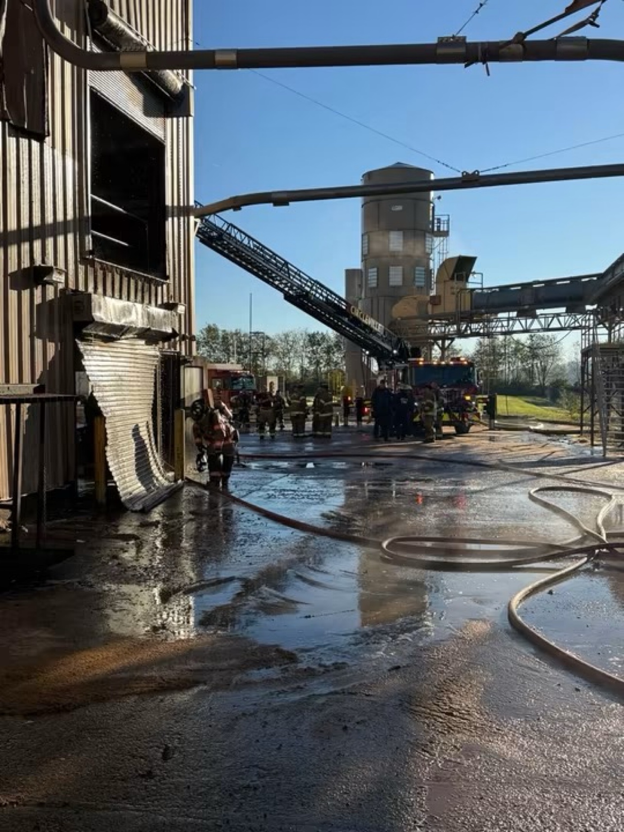 Firefighters work outside the plant, where a rollup door hangs damaged and hoses cover the wet lot.