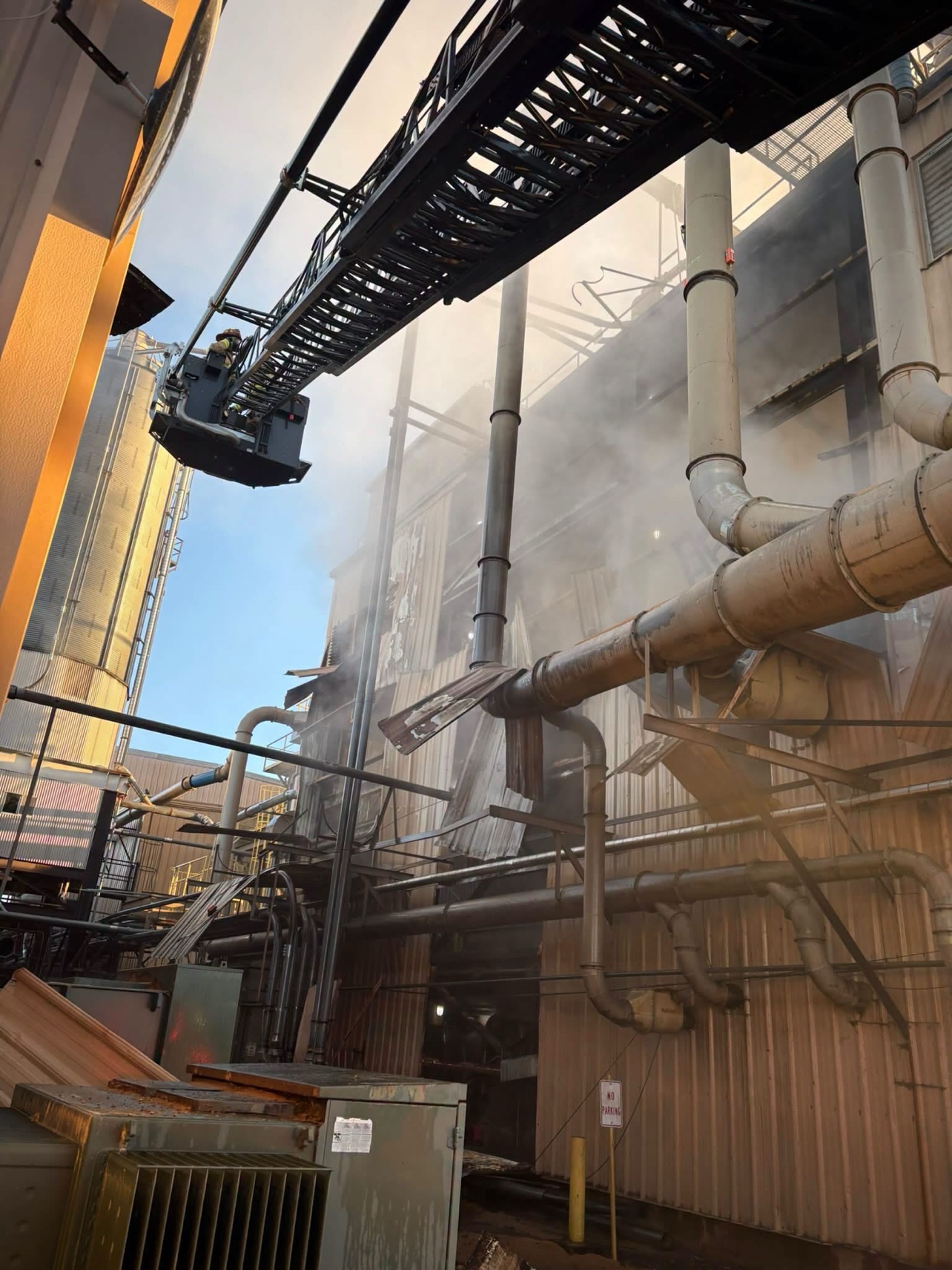 Smoke pours around the plant pipes and siding as the aerial ladder works above.