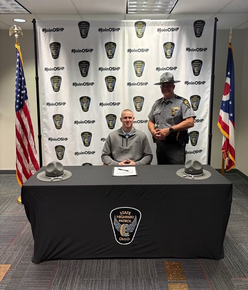 A cadet seated at the signing table with an Ohio State Highway Patrol trooper standing beside him